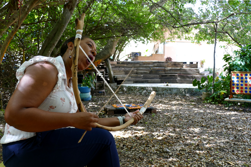 Sitting in the lush garden of Landhuis Bloemhof, Jennifer demonstrates how to play a traditional bowstring, an instrument former slaves brought with them from Africa.